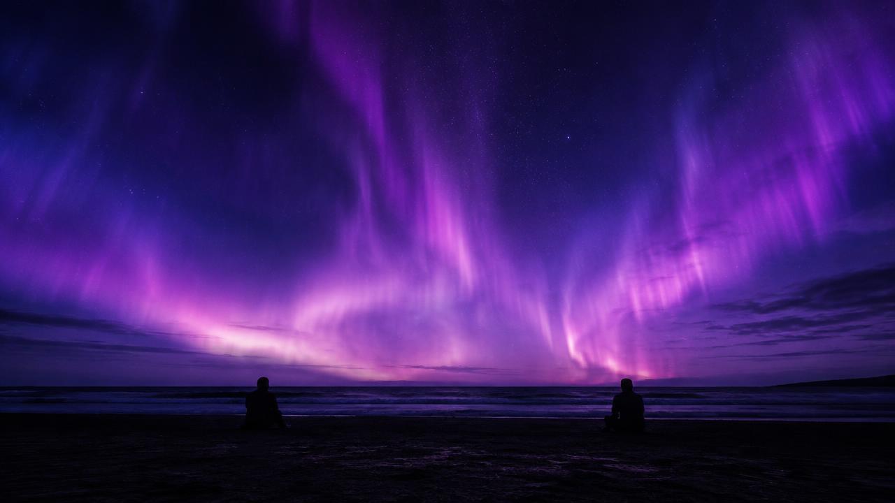 Two silhouettes sitting apart on a dark beach beneath a violet aurora